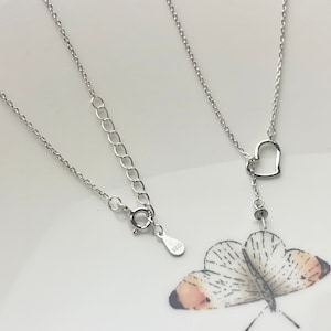 May include: A delicate silver necklace featuring a heart-shaped pendant. The necklace has a fine chain and an adjustable clasp. The pendant hangs from a vertical chain with a small, round charm. The necklace is displayed on a white surface.
