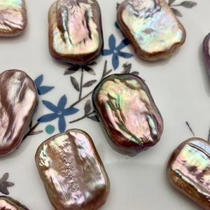 Puede incluir: Varias perlas rectangulares e iridiscentes. Las perlas presentan una superficie brillante con tonos de rosa, morado y verde. Se muestran sobre un plato blanco adornado con motivos florales azules. Ideales para la creación de joyas.