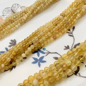 Puede incluir: Una hebra de cuentas de ópalo amarillo, cada cuenta es redonda y facetada. Las cuentas están enhebradas en un hilo transparente y se muestran sobre una superficie blanca con un patrón floral.