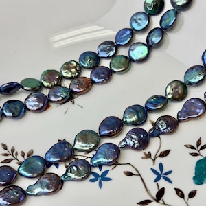 May include: Strands of iridescent, coin-shaped pearls in shades of blue, green, and purple. The pearls are arranged in rows, showcasing their unique, flat shape and lustrous surface. The background is a white plate with floral designs.