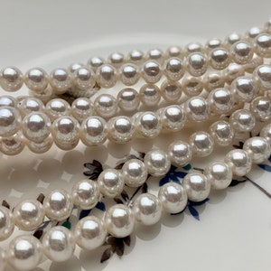 May include: A strand of white pearl beads, arranged in a layered pattern, on a white surface with a floral design.