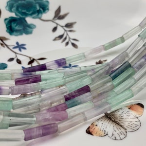 May include: A strand of light green, purple, and clear faceted gemstone beads. The beads are shaped like long, thin rectangles. The beads are arranged in a loose pile on a white surface.