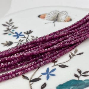May include: A strand of deep purple square-shaped gemstone beads. The beads are small and uniform in size and shape.