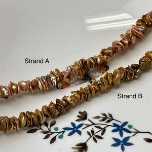 May include: Two strands of iridescent, chip-shaped beads. Strand A is a mix of pink and silver, while Strand B is golden brown. The strands are labeled on a white plate with floral accents. The beads appear to be natural.