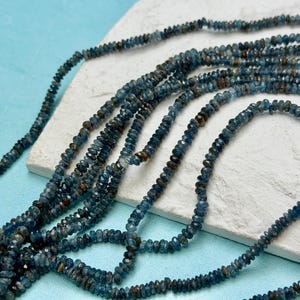 May include: Strands of blue-gray gemstone beads are displayed on a white surface against a light blue backdrop. The beads have a faceted cut, creating a sparkling effect. The beads are strung together, forming multiple necklaces.