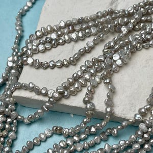 May include: A strand of white freshwater pearls with an irregular, slightly elongated shape. The pearls are strung on a clear thread and are arranged in a loose, flowing pattern.