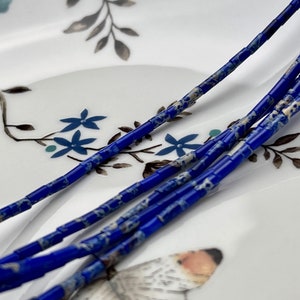May include: Close-up of a strand of blue and white gemstone beads. The beads are cylindrical and have a marbled pattern. The beads are arranged in a line on a white surface with a floral design.