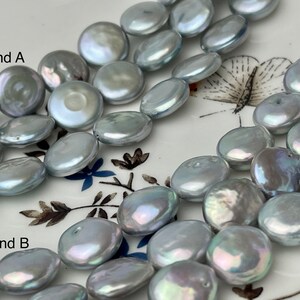 Puede incluir: Dos hebras de perlas de agua dulce grises. Las perlas son planas y redondas, con un ligero brillo iridiscente. Las hebras están etiquetadas como "Strand A" y "Strand B".