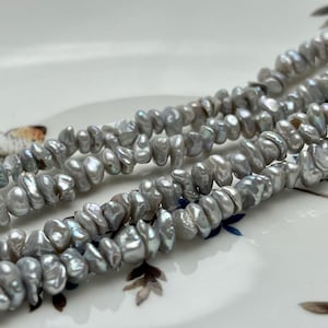 Puede incluir: Una hebra de perlas de agua dulce grises, con una forma irregular, ligeramente alargada. Las perlas están enhebradas en un hilo transparente.