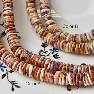 Puede incluir: Dos hebras de cuentas de concha, una con un patrón marrón y blanco etiquetada como "Color A" y la otra con un patrón marrón rojizo y blanco más pronunciado etiquetada como "Color B".