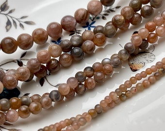 May include: A close-up of three strands of sunstone beads. The beads are round and have a variety of shades of brown, orange, and gold. The beads are arranged in a loose pile on a white surface.