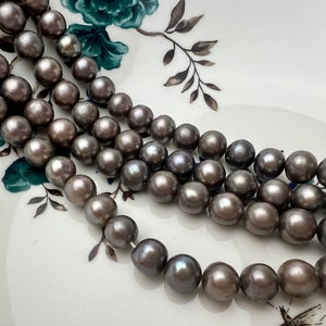 May include: A strand of gray pearls, with a slight iridescent sheen, arranged in a loose pile on a white surface with a floral pattern.