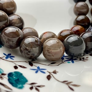 Puede incluir: Una colección de cuentas de piedras preciosas pulidas, redondas y de color marrón oscuro. Las cuentas tienen una superficie reflectante y diferentes tonos de marrón y negro. Se exhiben en un plato blanco con un diseño floral en verde azulado y azul.