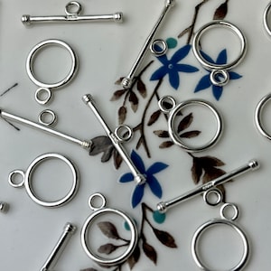 Puede incluir: Primer plano de cierres de palanca plateados con anillos redondos. Los cierres están esparcidos sobre una superficie blanca con un patrón floral.