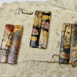 May include: Six rectangular stone pendants arranged in three pairs. Each pendant features a polished surface with unique natural patterns in shades of tan, brown, black, and muted red. Each piece has a small drilled hole at the top for hanging.