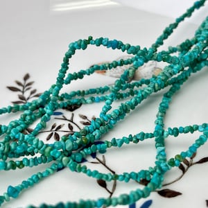 Puede incluir: Una hebra de cuentas de turquesa en forma de chip, de color azul verdoso claro, dispuestas en un montón suelto sobre una superficie blanca con un patrón floral.