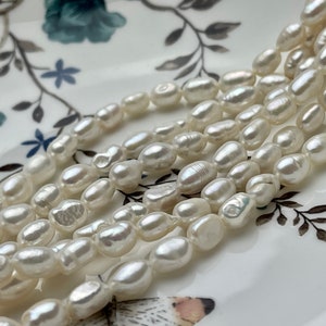 May include: A strand of white freshwater pearls, with a slightly irregular shape, arranged in a loose pile on a white plate with a floral pattern.