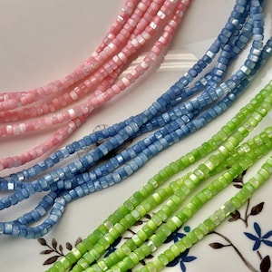 May include: Close-up of several strands of square-shaped beads in shades of pink, blue, and green. The beads are strung together, creating a textured and colourful display. The beads are likely used for jewellery making.