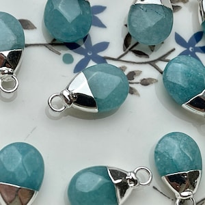 May include: A group of teal blue gemstone pendants with silver caps and jump rings. The pendants are on a white surface with a floral pattern.
