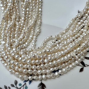 May include: A strand of white freshwater pearls, with a slightly irregular shape, arranged in a pile on a white plate with floral designs.