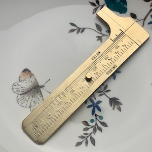 May include: A brass ruler with markings in inches and centimeters. The ruler is on a white surface with a floral pattern.