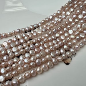 May include: A strand of pale pink freshwater pearls, with a slightly irregular shape, arranged in a loose pile on a white surface.