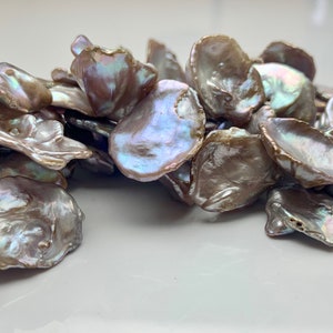 May include: A pile of iridescent, light brown, and gray baroque pearls. The pearls are irregularly shaped and have a natural, organic look.