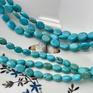 May include: Close-up of several strands of turquoise gemstone beads. The beads are oval-shaped and a vibrant shade of blue, with some showing natural variations. The beads are displayed on a white surface with a floral and butterfly design.