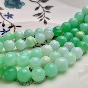 May include: A strand of green and white agate beads. The beads are round and have a smooth, polished finish. The beads are arranged in a gradient, with the green beads at one end and the white beads at the other.