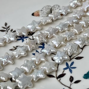 May include: A strand of white pearl beads shaped like stars. The beads are arranged in a row and are strung on a white thread. The beads are iridescent and have a slightly irregular shape.