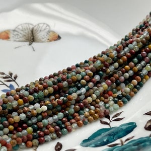 May include: A strand of multi-colored gemstone beads. The beads are small and round, and they are arranged in a long line. The beads are a mix of green, brown, red, and white.