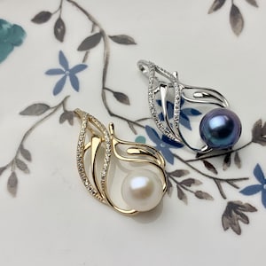Puede incluir: Dos delicados colgantes de perlas con diseño de hoja. Un colgante presenta una perla blanca y el otro una perla azul oscuro. Ambos colgantes están adornados con pequeñas piedras transparentes.