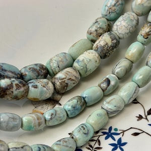 May include: Strands of turquoise-colored, oval-shaped beads with brown and white marbling. The beads are arranged in parallel rows, showcasing their unique patterns and textures. The beads are displayed on a white plate with a floral design.