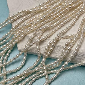 May include: Close-up of multiple strands of creamy white, oval-shaped pearls. The pearls are strung together, creating a delicate and elegant appearance. The background is a light blue surface with a white, textured surface.