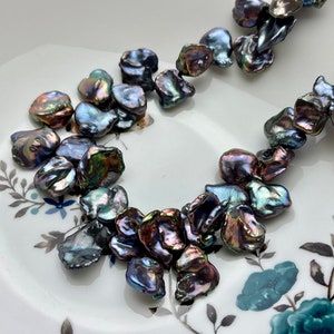 May include: A strand of iridescent black baroque pearls, with a variety of shapes and sizes, displayed on a white plate with a floral pattern.