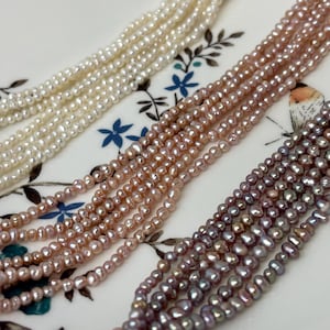 May include: Close-up of several strands of small, round pearls in shades of white, pink, and purple. The pearls are strung together, creating a delicate and elegant appearance. The strands are arranged on a decorative plate with floral patterns.
