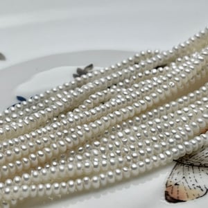 May include: A strand of white freshwater pearls, arranged in a fan shape on a white surface. The pearls are small and uniform in size.