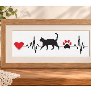 Könnte beinhalten: Ein rechteckig gerahmtes Kreuzstich-Stickbild mit einer schwarzen Herzschlaglinie auf weißem Grund. Das Design zeigt ein rotes Herz, die schwarze Silhouette einer laufenden Katze sowie einen schwarz-roten Pfotenabdruck in der Pulsline.