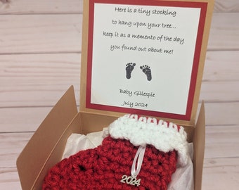 May include: A small red crocheted stocking with a white cuff and a gold 2024 charm. The stocking is in a brown box with white tissue paper lining. The box has a card inside with the text "Here is a tiny stocking to hang upon your tree... keep it as a memento of the day you found out about me! Baby Gillespie July 2024" and two footprints.