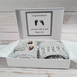 May include: A baby gift set in a white box. The set includes a gray crocheted hat and booties with wooden button closures. The box has a card that reads "Congratulations!" and "We can't wait to meet Baby Hall!" with footprints.