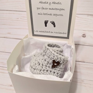 May include: A white gift box with a gray card inside. The card has a message written in Spanish that says "Abuela y Abuelo, por favor mantengan mis botines seguros. Los necesitar&eacute; en Noviembre." The box contains a pair of gray crocheted baby booties with a brown heart button.
