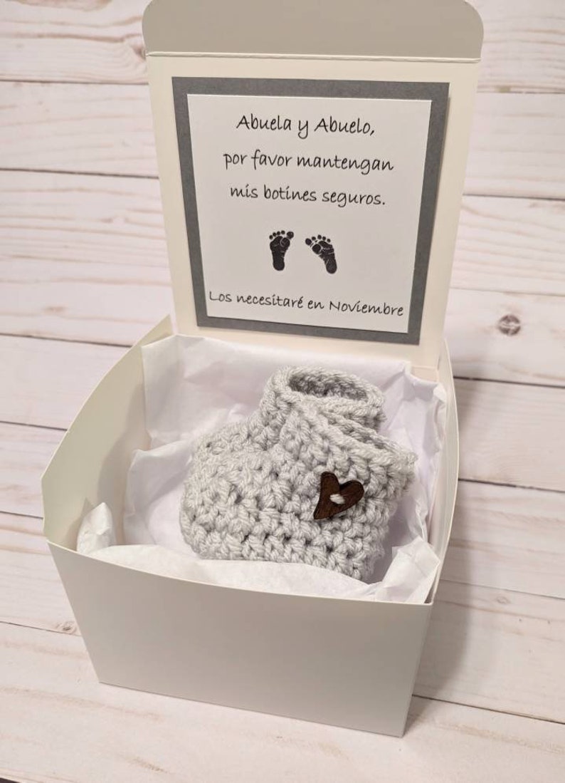 May include: A white gift box with a gray card inside. The card has a message written in Spanish that says "Abuela y Abuelo, por favor mantengan mis botines seguros. Los necesitar&eacute; en Noviembre." The box contains a pair of gray crocheted baby booties with a brown heart button.