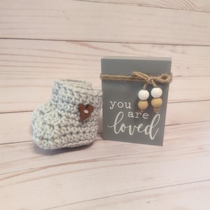 May include: Gray crocheted baby booties with a brown heart button and a gray wooden sign with the text "you are loved" and white and tan beads.