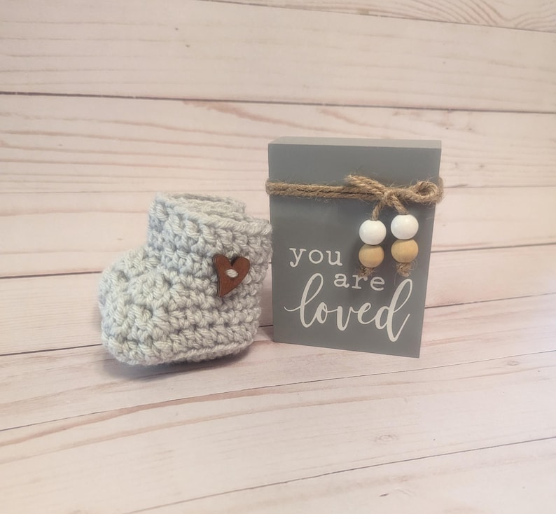 May include: Gray crocheted baby booties with a brown heart button and a gray wooden sign with the text "you are loved" and white and tan beads.