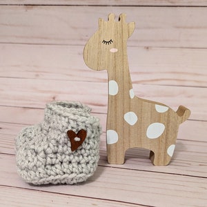 May include: A gray crocheted baby bootie with a brown heart-shaped button. The bootie is next to a wooden giraffe toy with white polka dots.