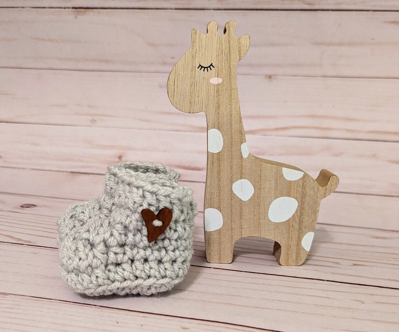 May include: A gray crocheted baby bootie with a brown heart-shaped button. The bootie is next to a wooden giraffe toy with white polka dots.