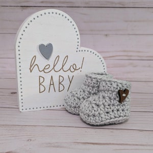 May include: Gray crocheted baby booties with a wooden heart button. A white heart-shaped sign with the text "hello! BABY" is in the background.