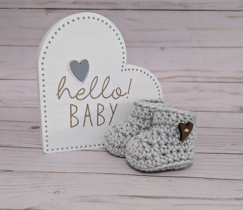 May include: Gray crocheted baby booties with a wooden heart button. A white heart-shaped sign with the text "hello! BABY" is in the background.