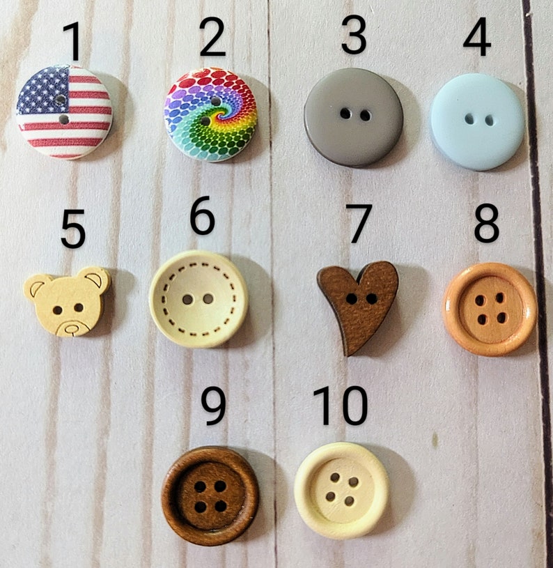 May include: Ten different wooden buttons with various designs, including an American flag, a rainbow swirl, a gray circle, a light blue circle, a teddy bear, a heart, a brown circle with four holes, a beige circle with two holes, a brown circle with four holes, and a beige circle with six holes.