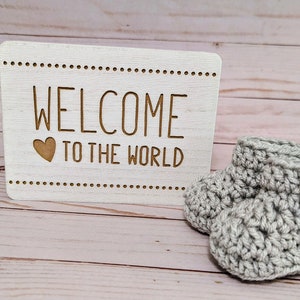 May include: A white wooden sign with the text "Welcome to the world" and a small heart.  A pair of gray crocheted baby booties with a wooden heart button.